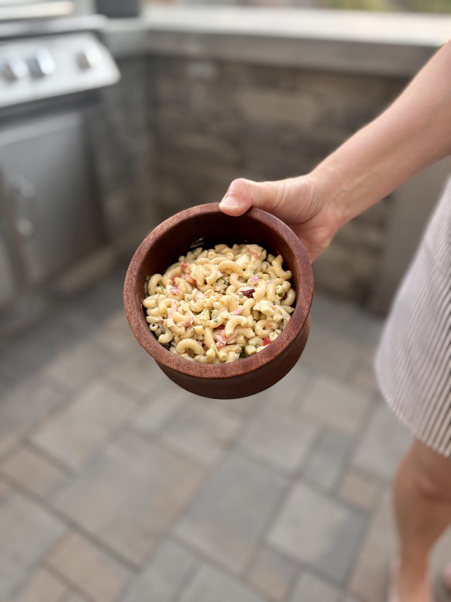 A hand holding a small terracotta bowl of creamy dressed macaroni salad, standing beside an outdoor grill on a paver patio.