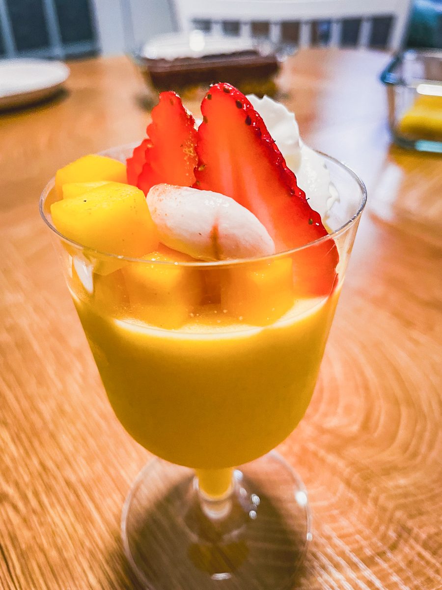 A stemmed glass of bright orange mango panna cotta topped with mango slices, cream and a strawberry, sitting on a wooden table.