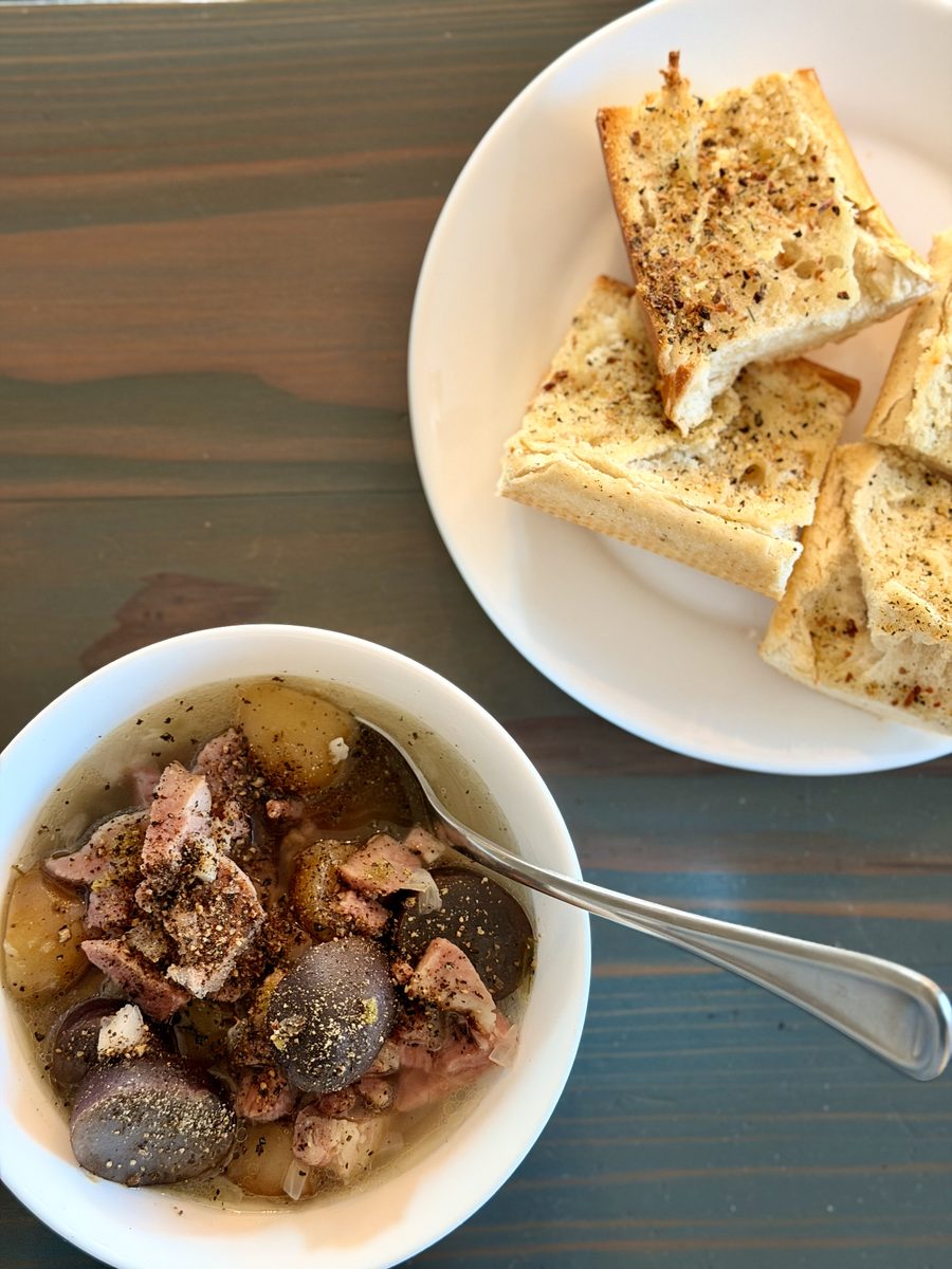 An overhead shot of a white bowl of brothy stew with chunks of meat and gold and purple potatoes, a spoon in it, beside a white pl