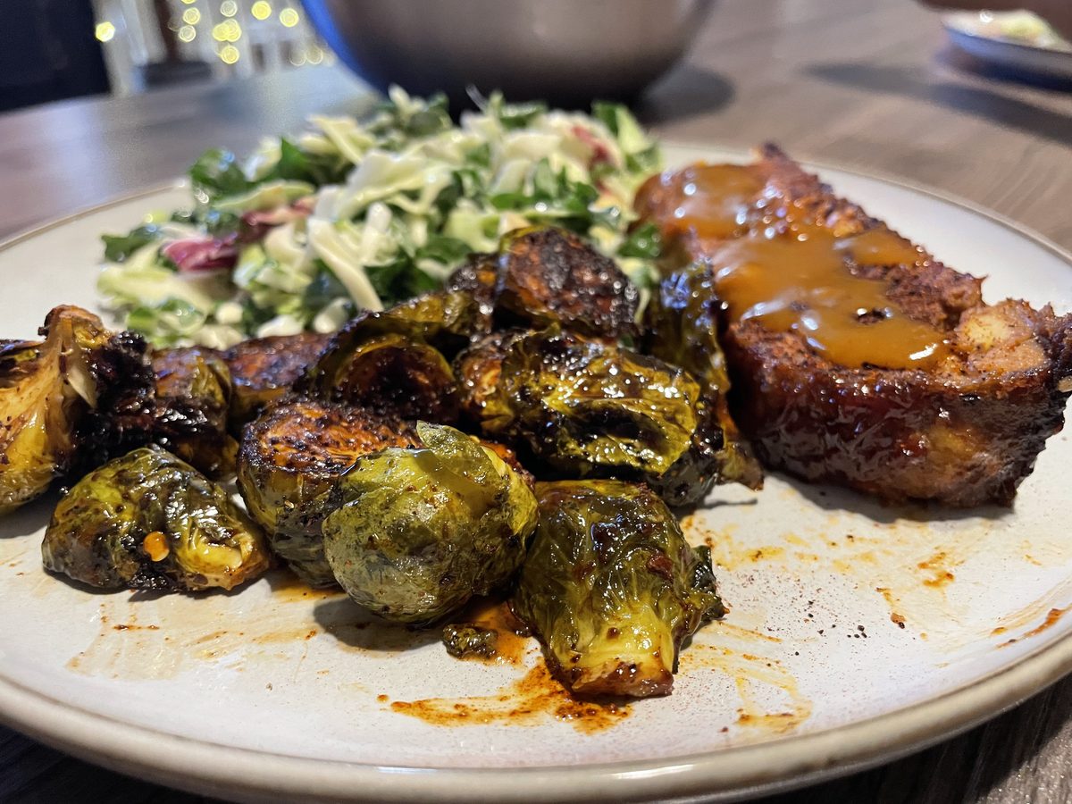 A dinner plate holding a slab of glazed meatloaf with gravy, charred roasted brussels sprouts and a shredded cabbage salad, on a d