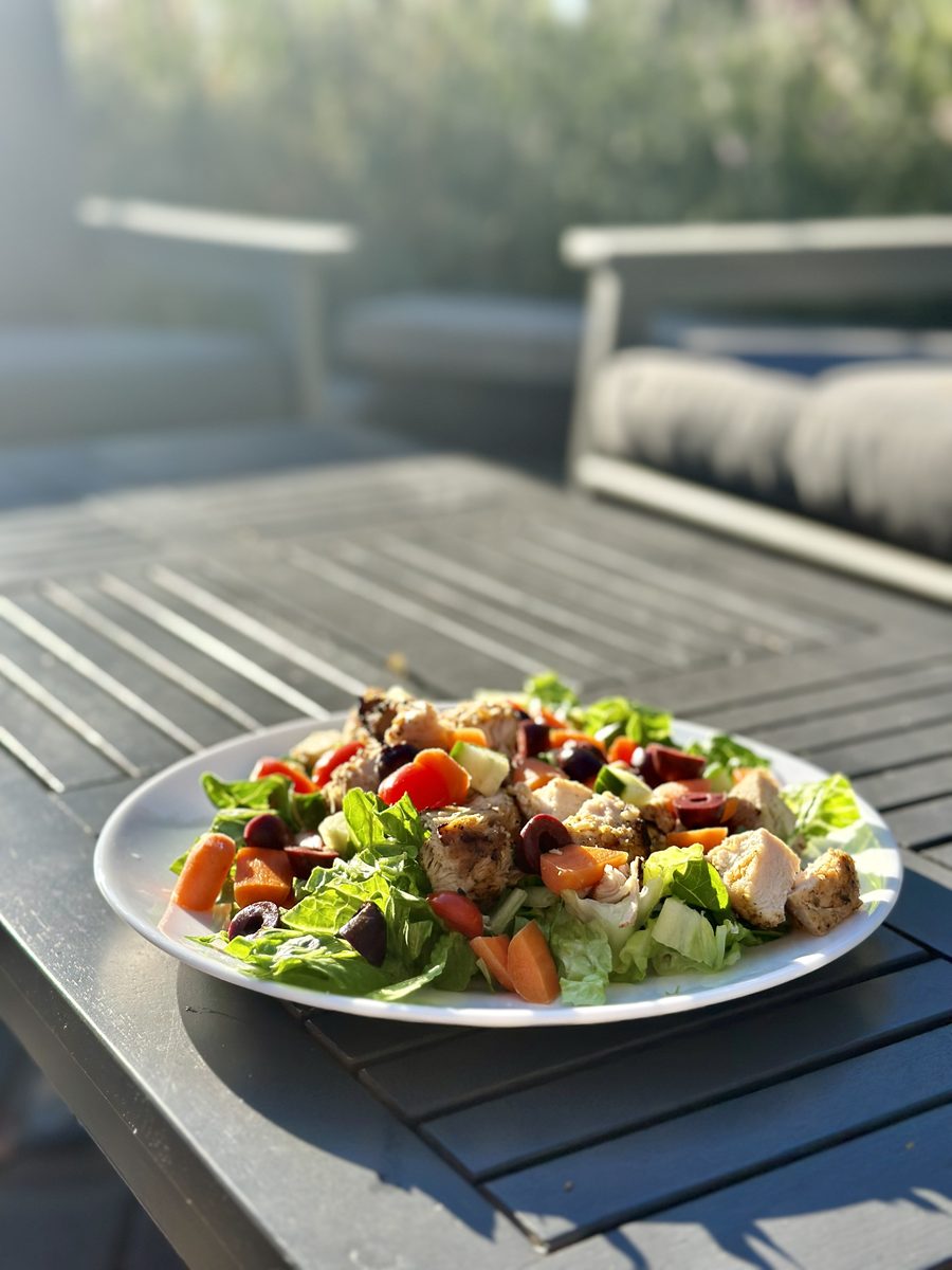 A white plate of Mediterranean salad with lettuce, tomato, olives, feta and grilled chicken pieces sitting on a gray slatted patio
