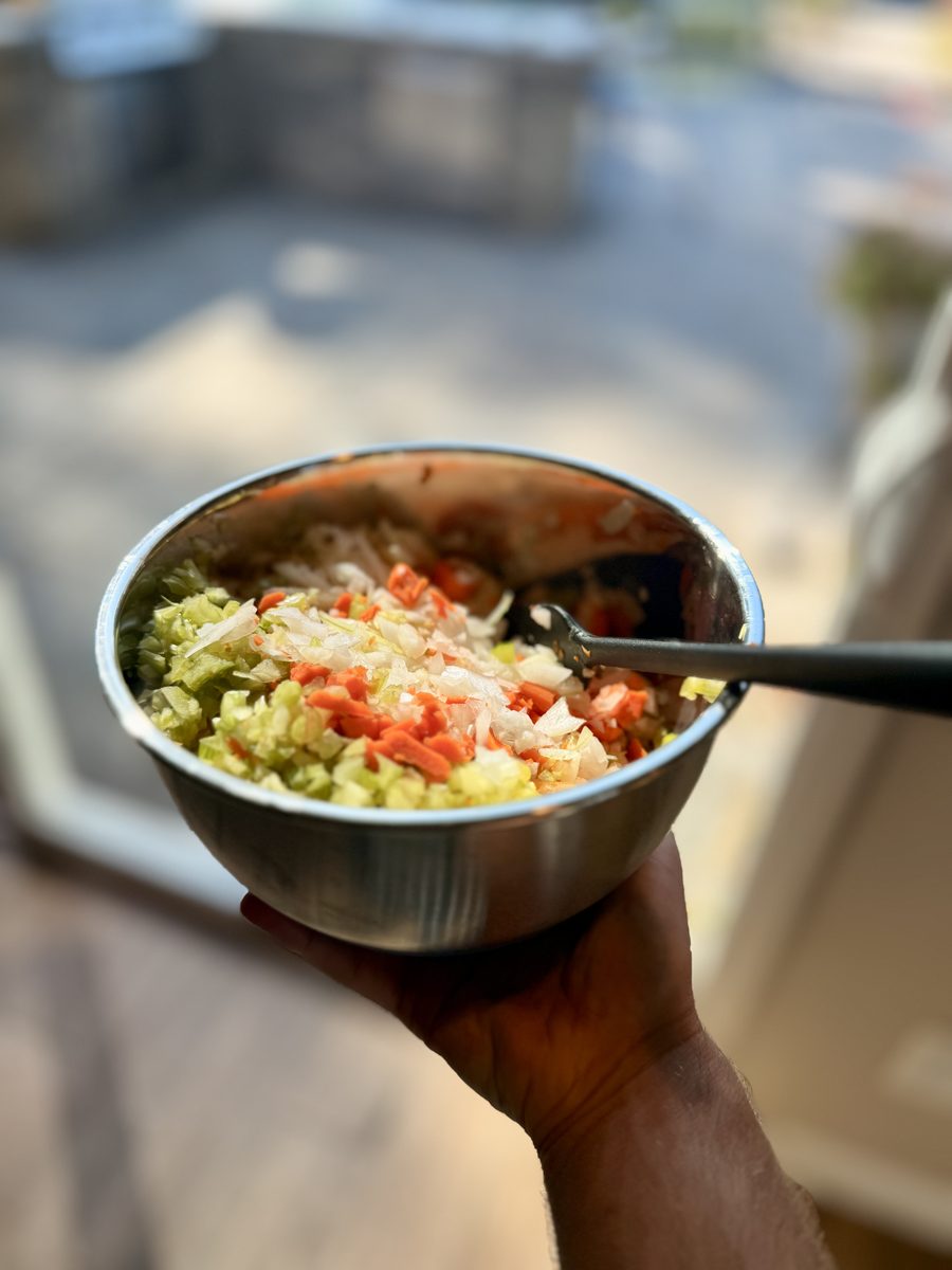 A hand holding a small stainless steel bowl of chopped vegetable salad with tomato, cucumber, onion and crumbled cheese and a spoo