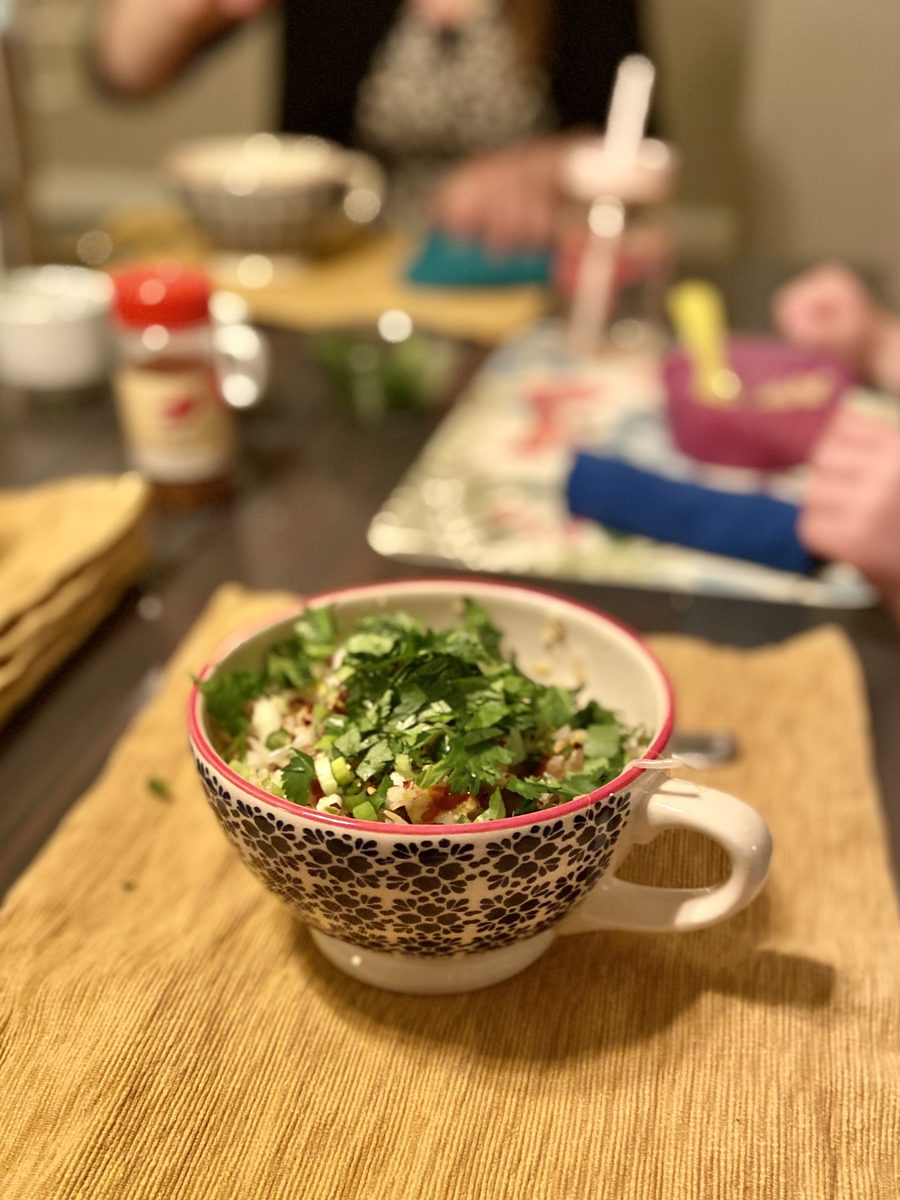A patterned ceramic mug of rice soup heaped with chopped cilantro on a woven placemat, with a dim blurred dinner table and people 
