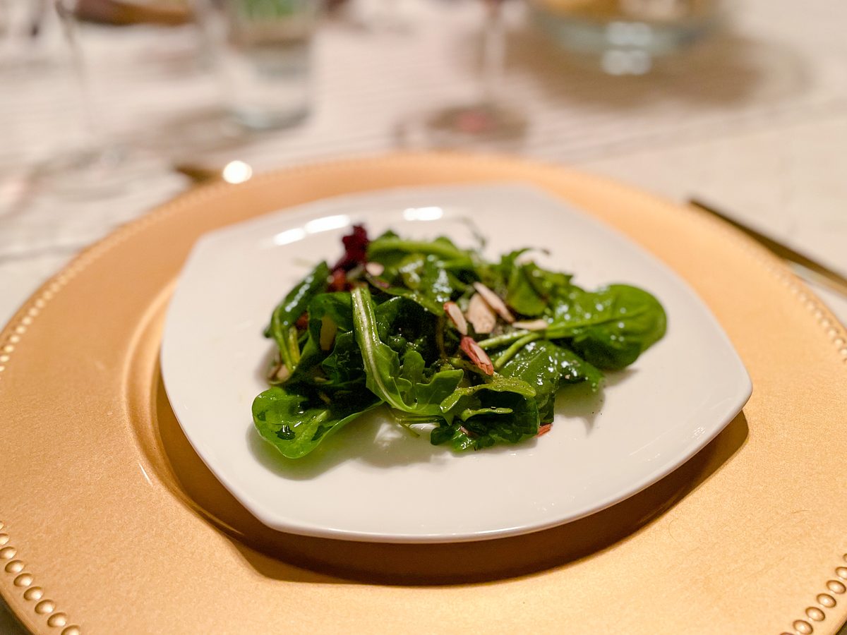 A small square white plate of mixed greens salad with sliced almonds and radicchio on a gold charger at a set dinner table, dim in