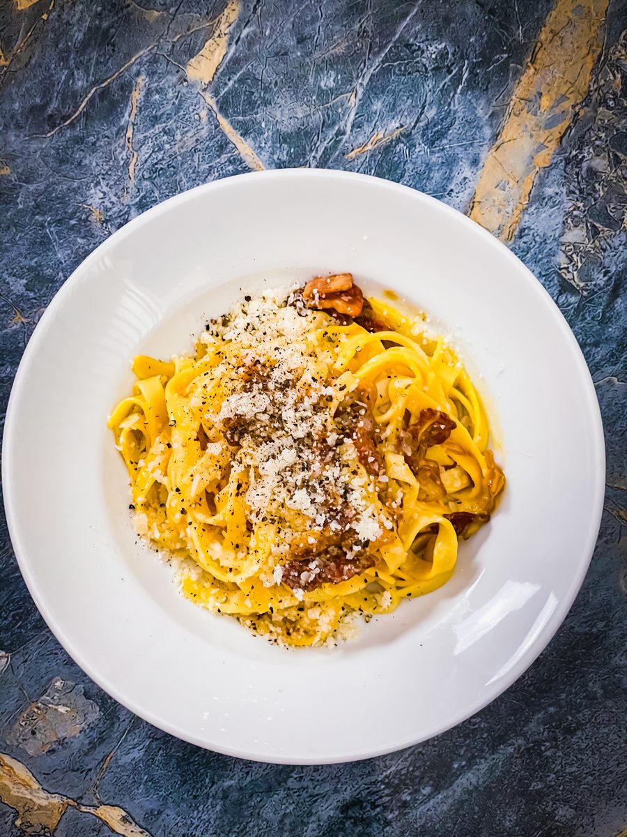 An overhead white bowl of creamy fettuccine with browned mushroom-like pieces, grated parmesan and cracked black pepper, on a blue