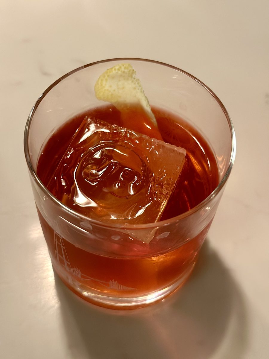 A rocks glass of deep red negroni over a large ice cube with an orange peel twist, on a white marble counter