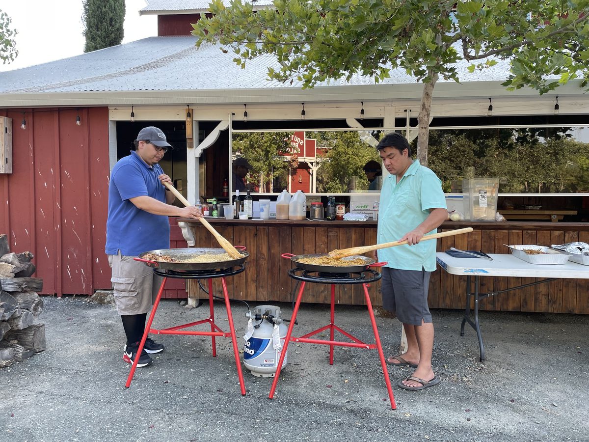 Two men stirring two large paella pans on red propane burner stands on a gravel driveway outside a red barn with an outdoor bar, c