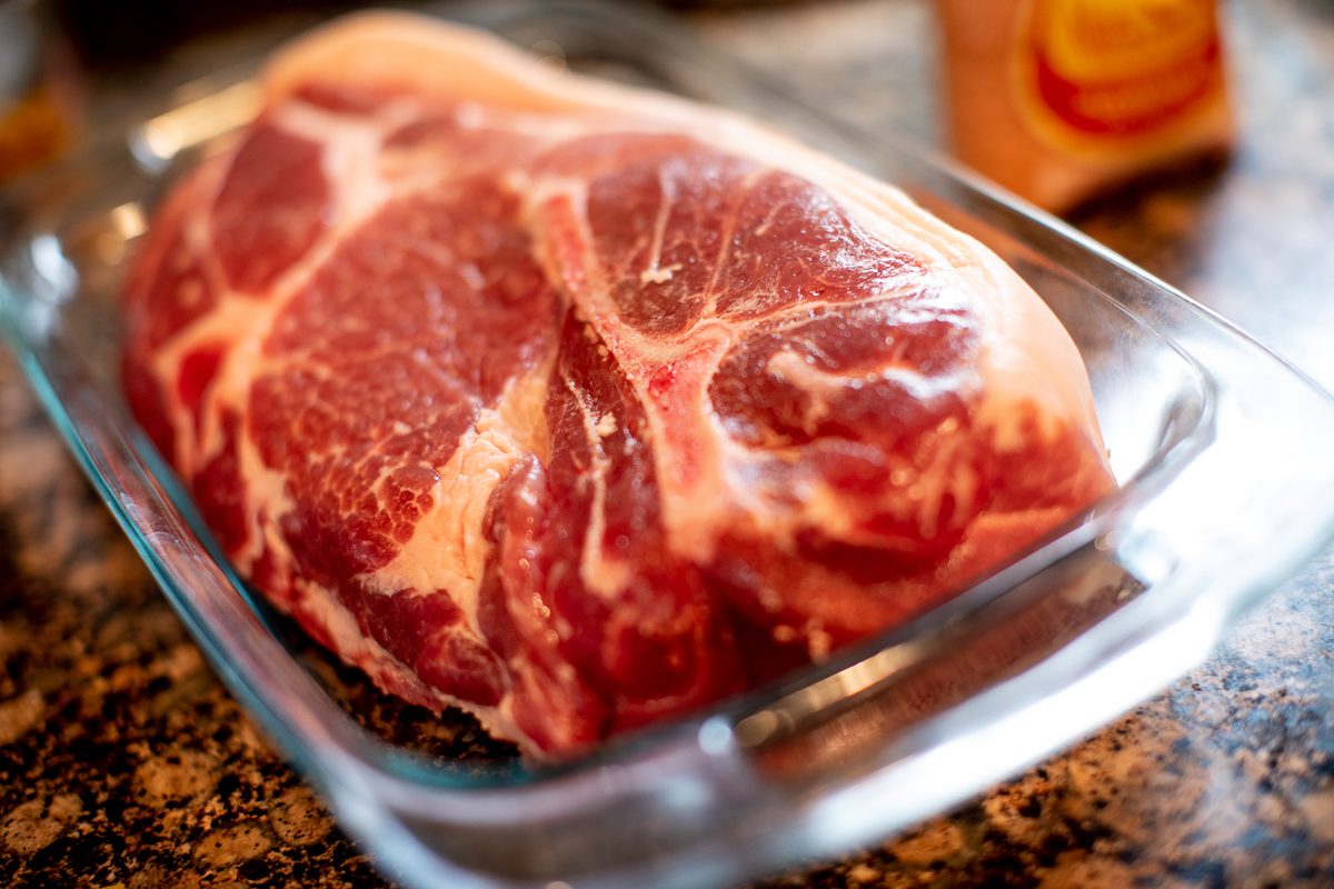 A large raw marbled pork shoulder roast sitting in a clear glass baking dish on a dark granite counter with blurred packages behin