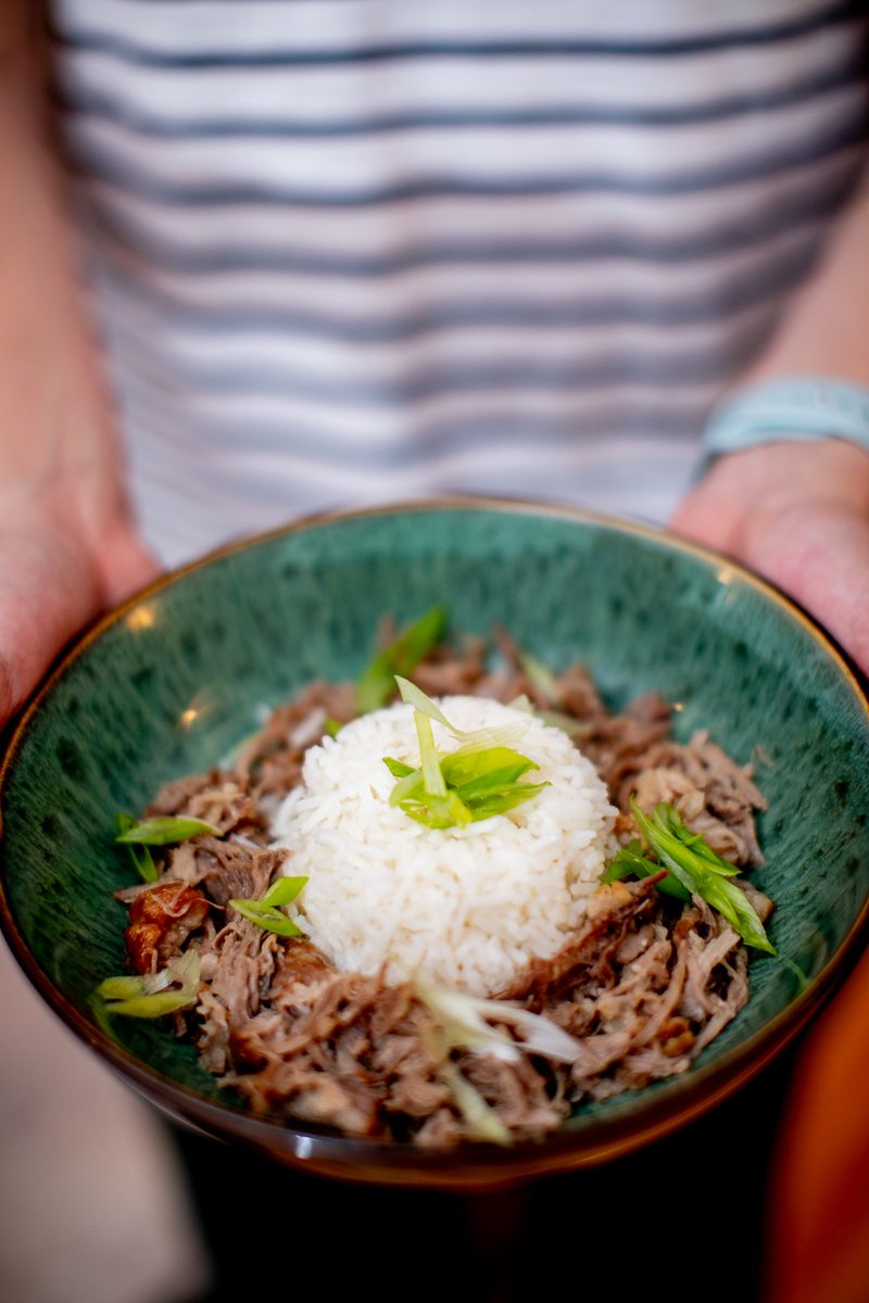 A person in a striped shirt holds a green speckled ceramic bowl of shredded kalua pork ringed around a mound of white rice with sl