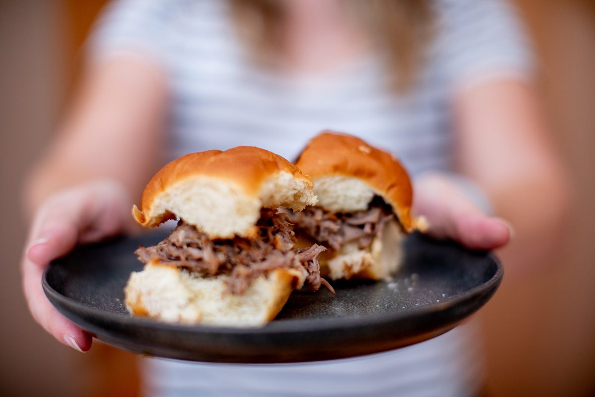 Hands hold a dark round plate with two pulled pork sliders on soft golden rolls; person in a striped tee blurred behind.