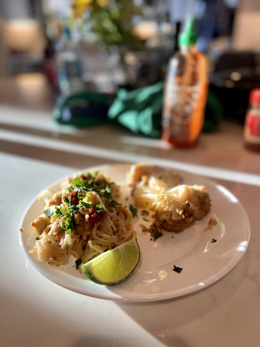 The same plate of pad thai noodles with a lime wedge and a breaded fried piece on a counter, shot wider with a cluttered kitchen b
