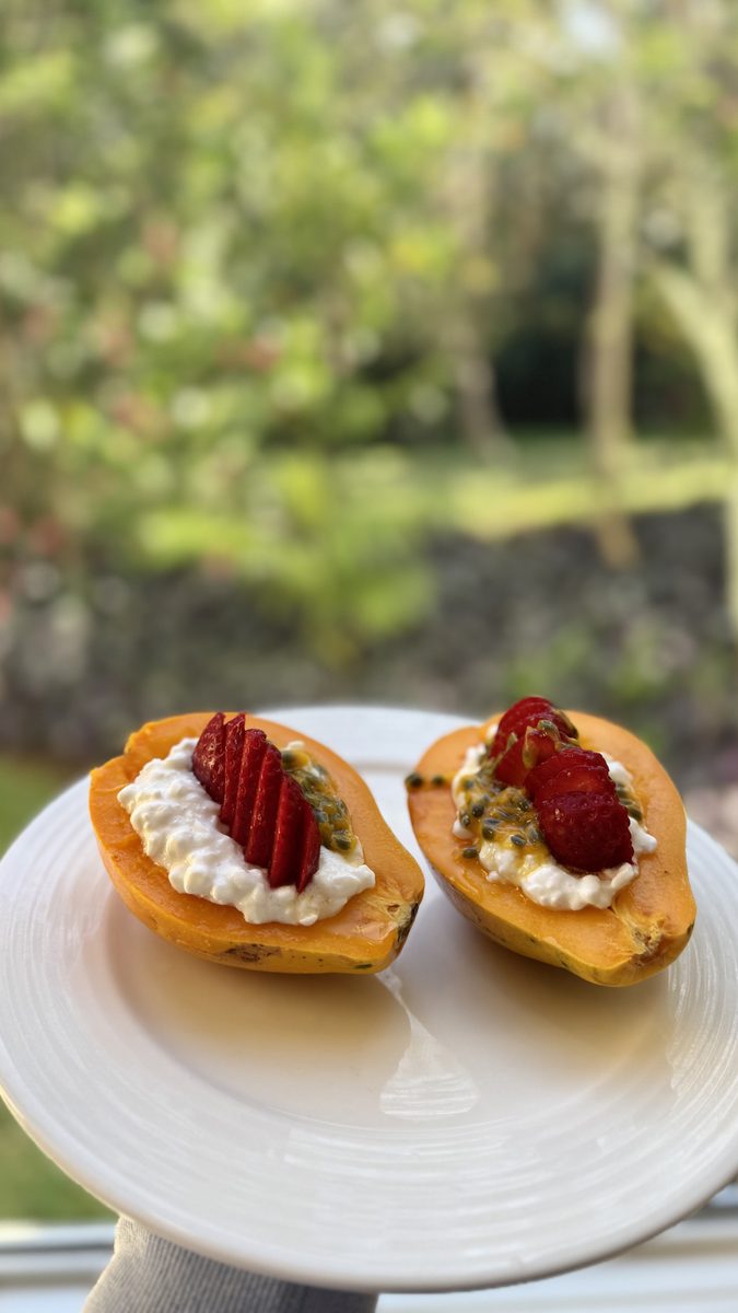 Two papaya halves filled with yogurt or cottage cheese and topped with sliced strawberries and seeds, on a white cake stand with a