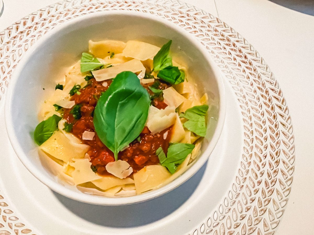 A white bowl of wide pappardelle noodles with meaty bolognese sauce, shaved parmesan and fresh basil leaves, on a white woven plac