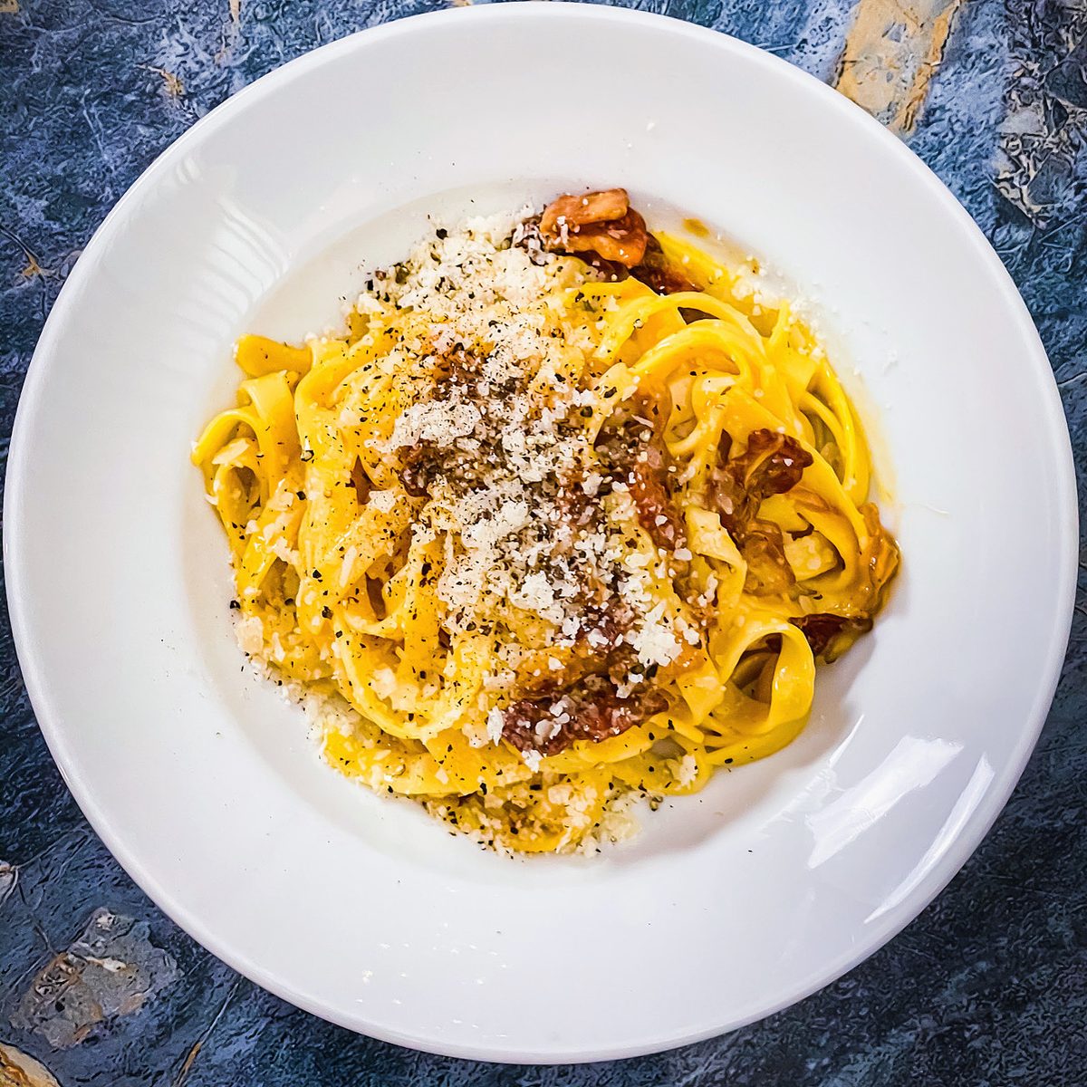 Overhead shot of a white shallow bowl of tagliatelle carbonara tossed in a glossy yellow egg sauce with browned pancetta pieces, g