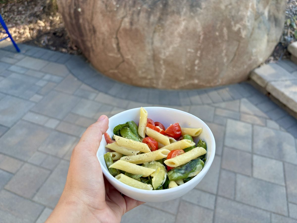 Two hands hold a white bowl of penne primavera with tomato, zucchini and greens outdoors over a paver patio.