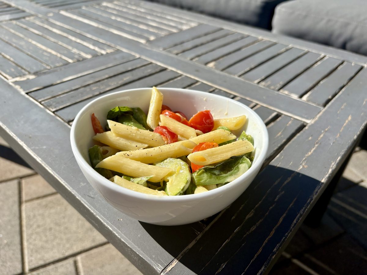 A white bowl of penne pasta with green vegetables and red pepper strips, sitting on a grey slatted metal patio table.