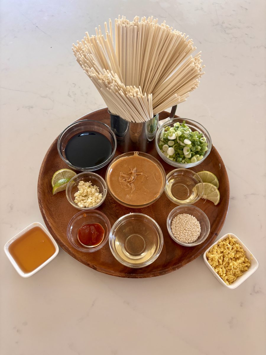 Overhead ingredient spread on white marble: a jar of dry wheat noodles standing upright behind a round wood board holding bowls of