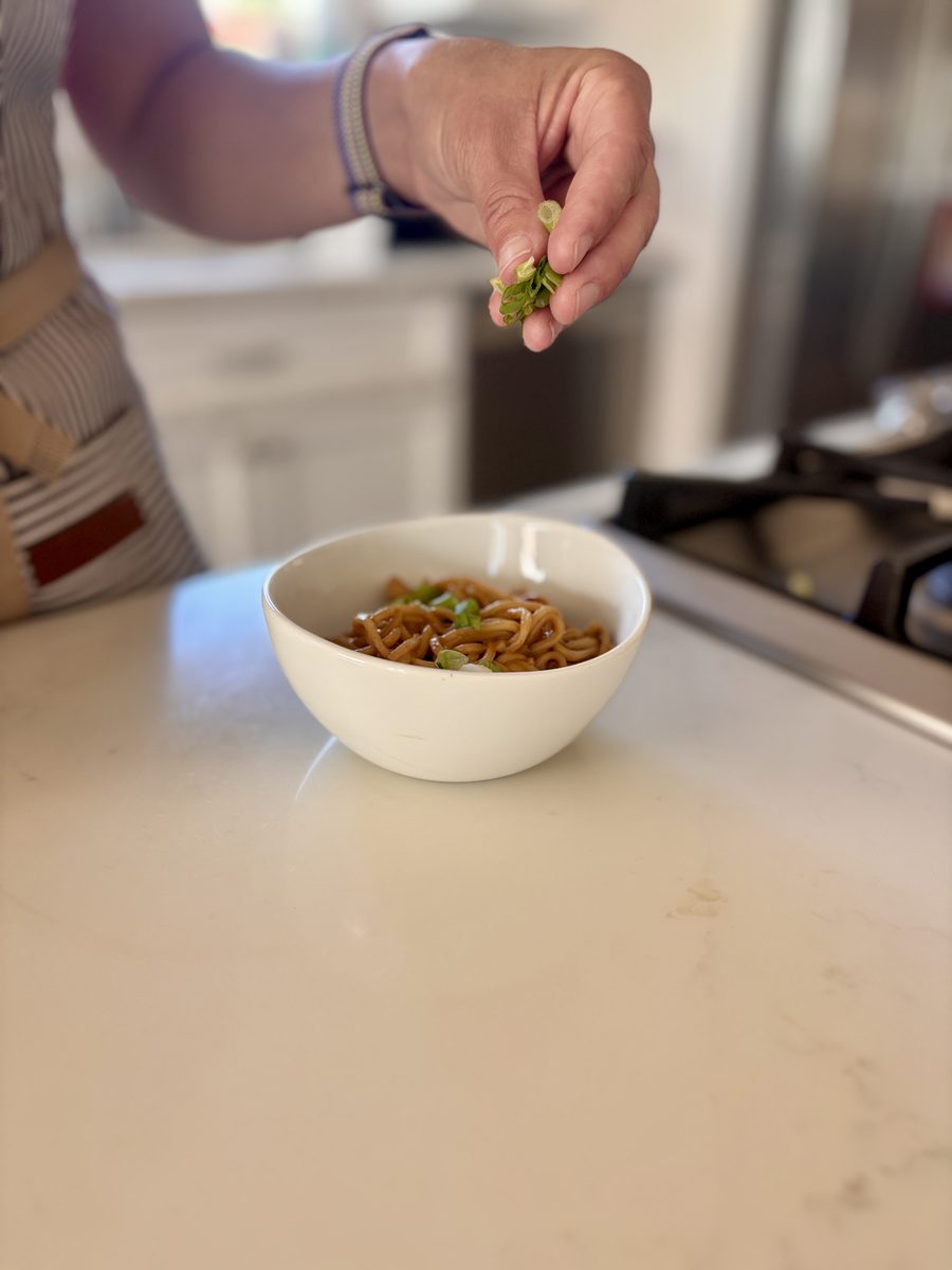 A small white bowl of peanut noodles sits on a pale kitchen counter while a hand above sprinkles sliced scallions over it.