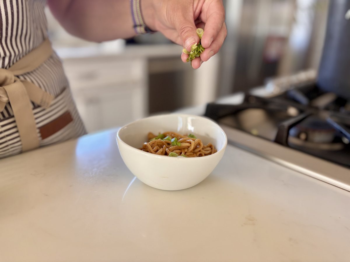 A white bowl of finished peanut noodles on a counter next to the range, with a hand dropping chopped scallions onto it from above.