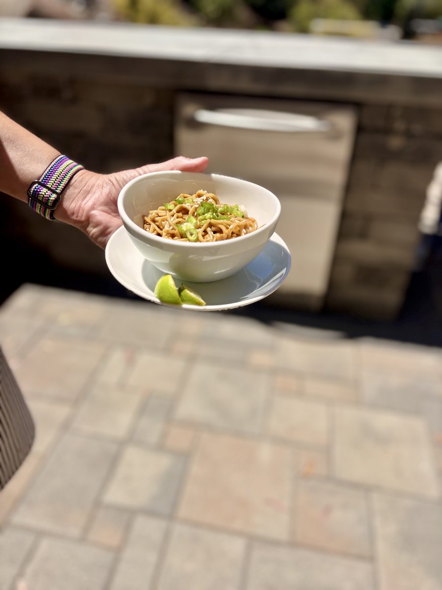 A hand holds out a white bowl of peanut noodles topped with scallions on a saucer with lime wedges, over a sunlit stone patio.