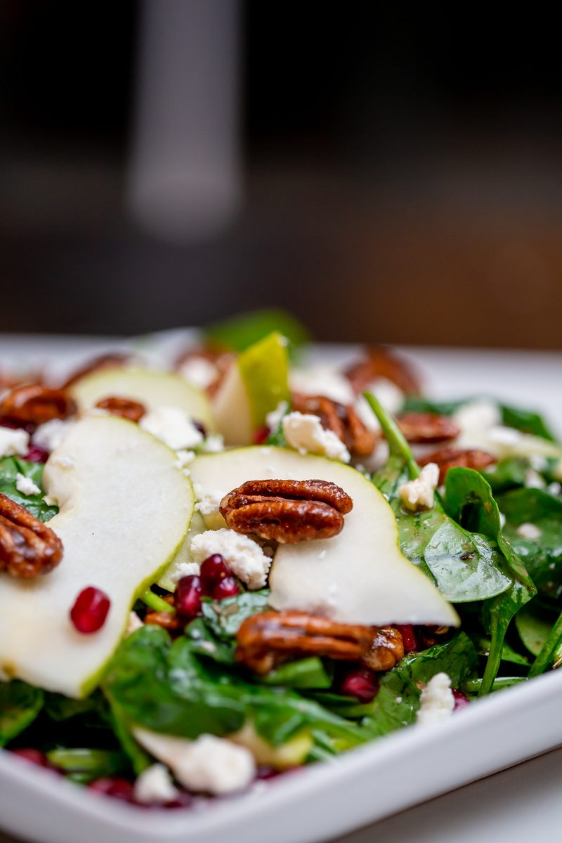 A white square plate of spinach salad with sliced pears, candied pecans, pomegranate arils and crumbled white cheese, photographed
