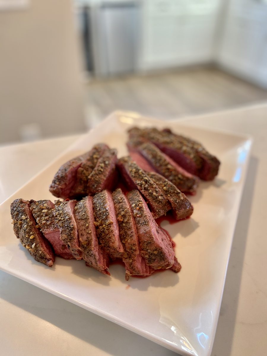 A white rectangular plate holding a pepper-crusted beef roast sliced into thick rare pink slices, on a light counter with a bright