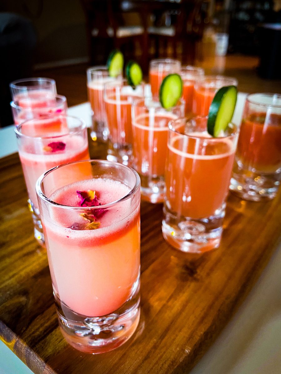 A wooden board of tall pink cocktails garnished with cucumber ribbons and edible flowers