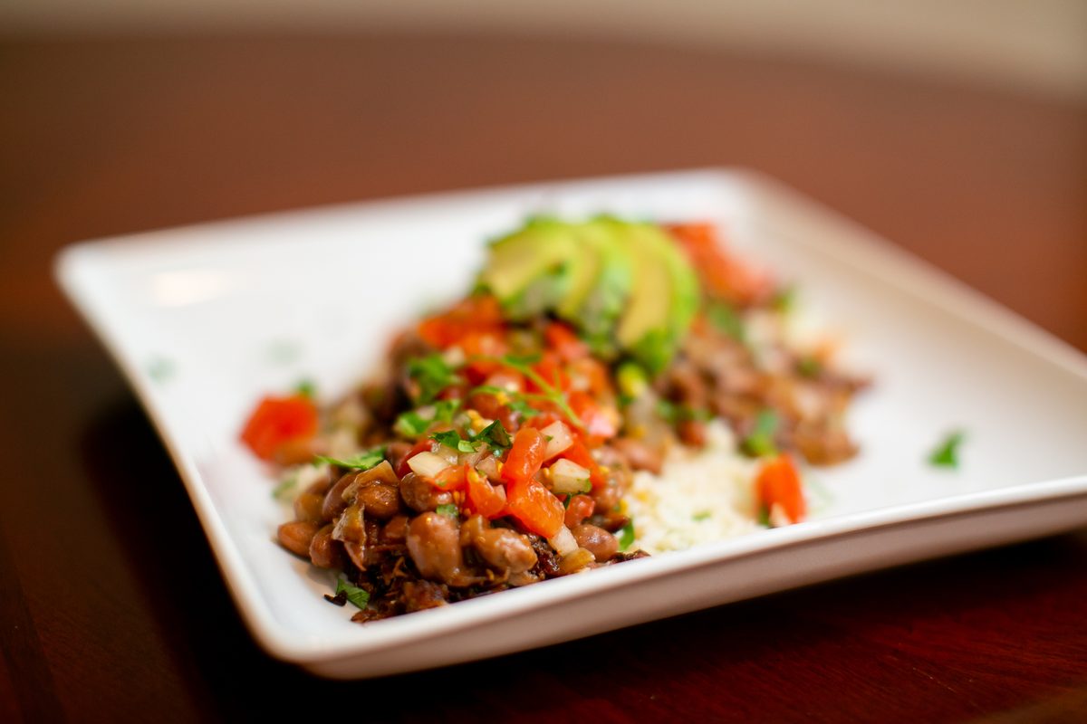 A white rectangular plate of pinto beans and cilantro-lime rice topped with diced tomato and avocado slices, shot at a low angle o