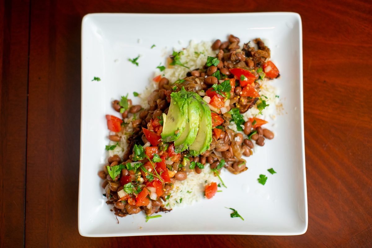 Overhead shot of a white square plate of pinto beans over white rice with pico de gallo, cilantro and fanned avocado slices, on a 