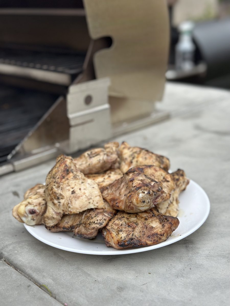 A white plate stacked with pieces of seasoned grilled chicken sitting on a counter beside an outdoor grill.