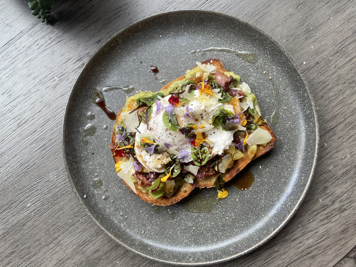 A thick slice of toast spread with mashed avocado and topped with a poached egg, micro greens, edible flower petals, shaved cheese