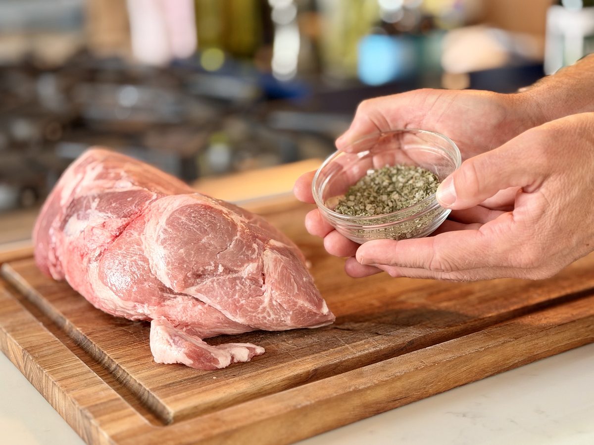A large raw pork shoulder roast sitting on a wooden cutting board while two hands hold a small glass bowl of dried herb rub beside