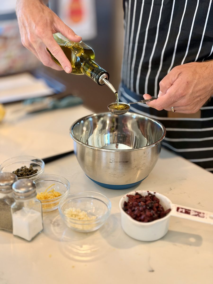 A person in a striped apron pours olive oil from a bottle into a stainless steel mixing bowl on a white counter, with small bowls