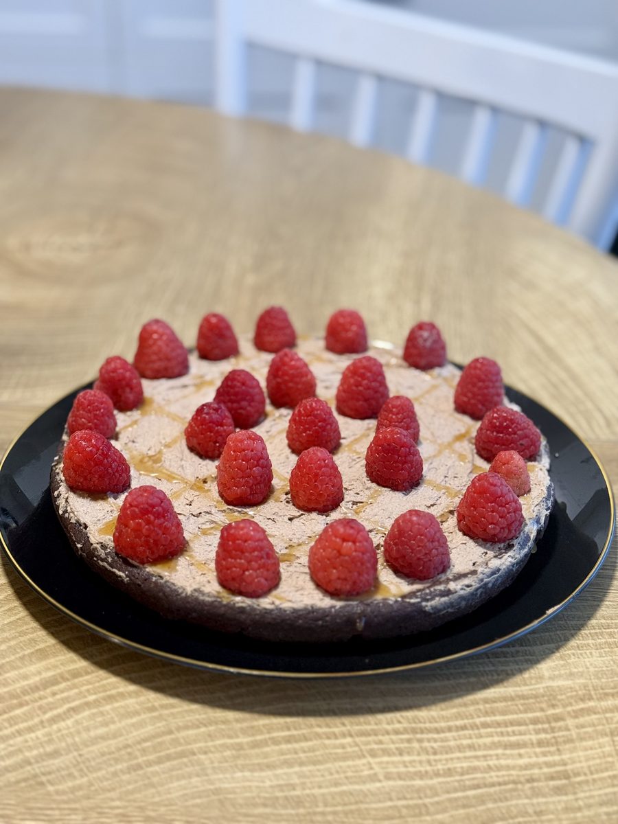 A whole round cake with a chocolate base and pale cream topping ringed with fresh raspberries, on a black plate on a light wood ta
