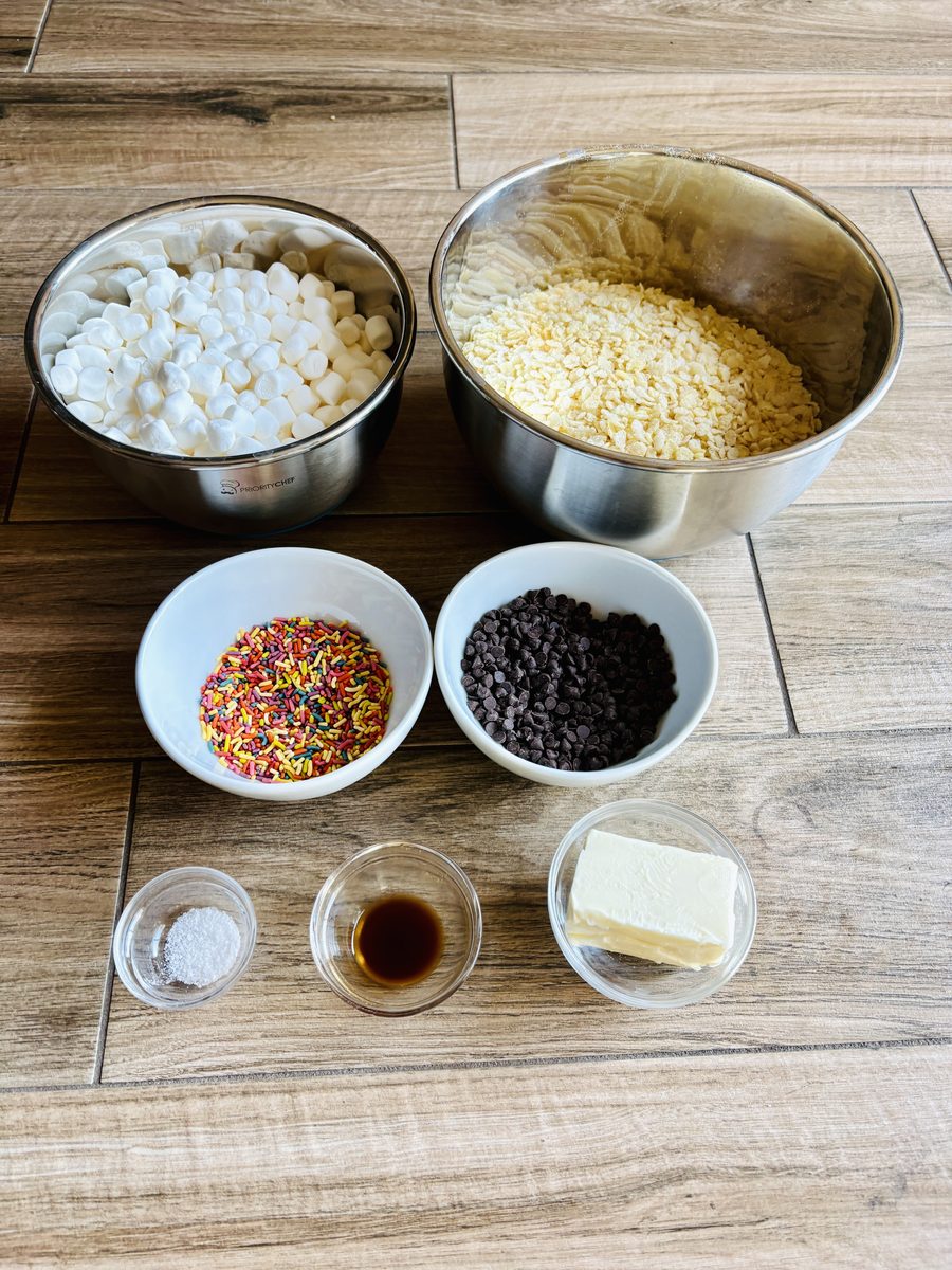 Overhead ingredient layout on a wood-plank surface: steel bowls of mini marshmallows and crisped rice cereal, white bowls of rainb
