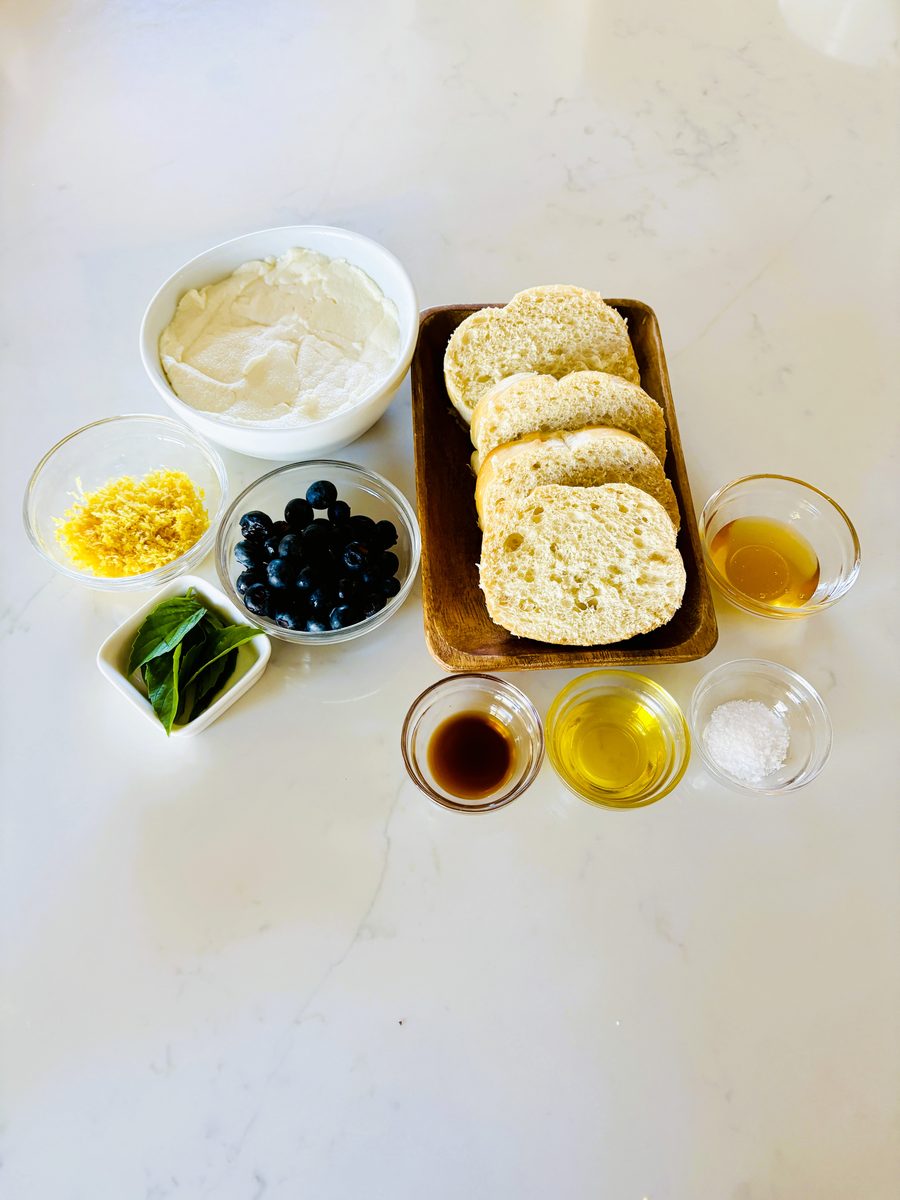 Overhead ingredient spread on a white counter: a bowl of ricotta, sliced baguette rounds on a wood tray, blueberries, lemon zest,