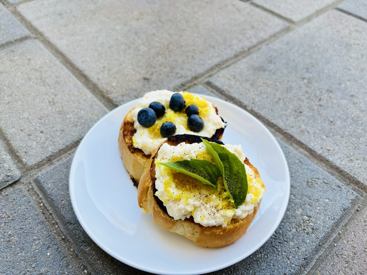 A small white plate on a paved patio with two open-faced toasts spread with soft white ricotta, one topped with blueberries and a 