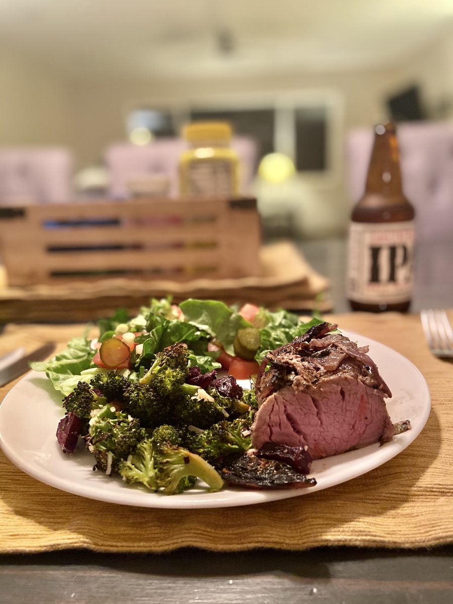 A white plate on a wood board holding a thick slice of medium-rare roast beef, charred roasted broccoli and a green salad with tom