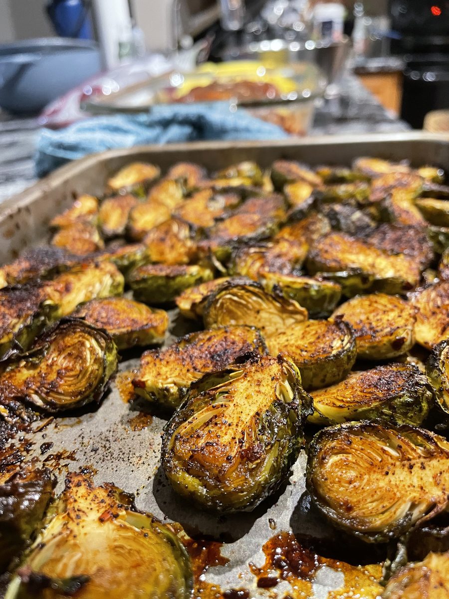 Close overhead of deeply caramelized halved brussels sprouts spread on a sheet pan, with a cluttered kitchen counter visible at th
