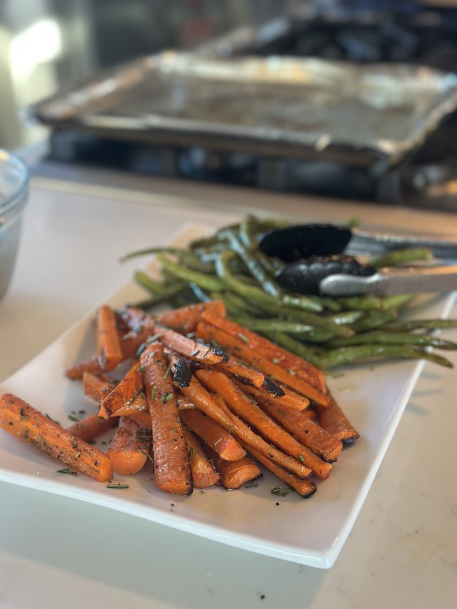 A white square platter of roasted carrot sticks alongside roasted green beans, with a used sheet pan and dark oven visible behind