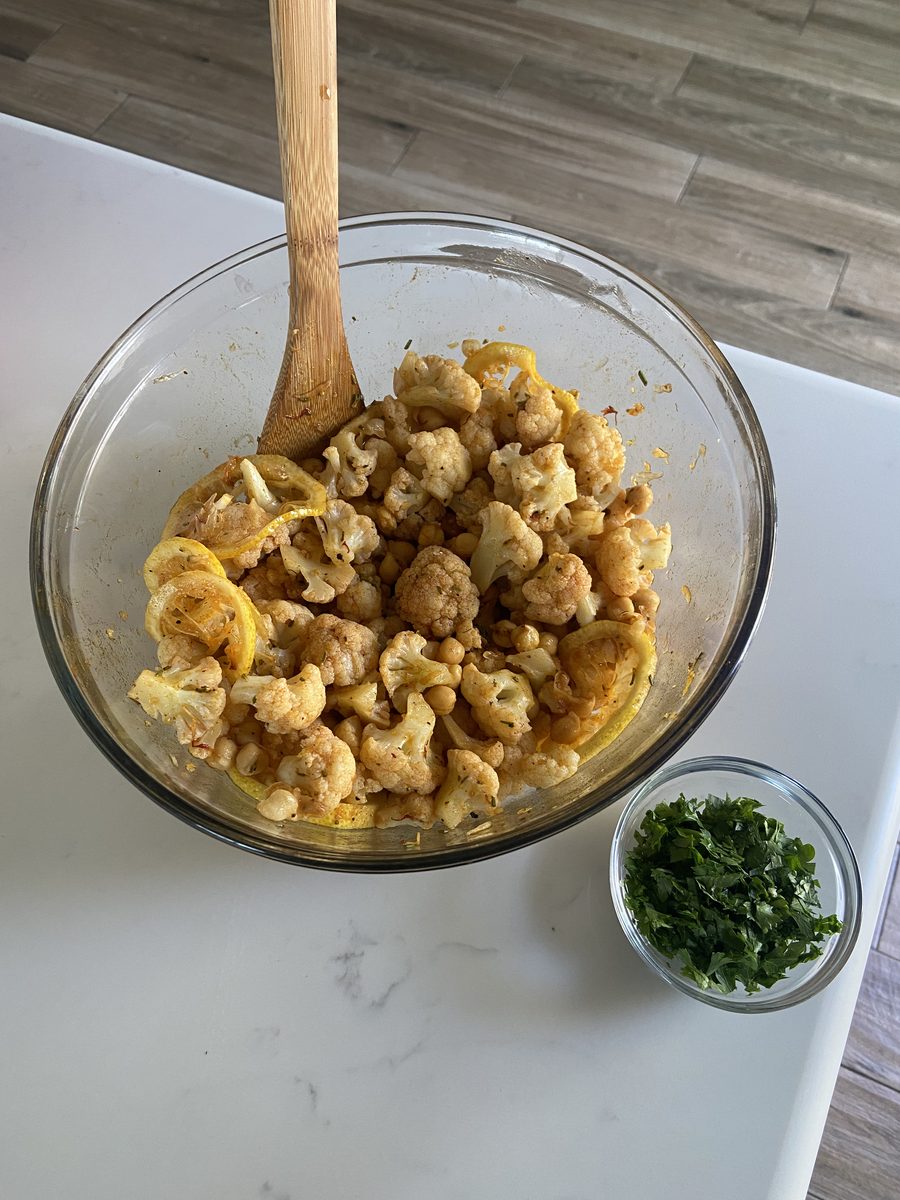 A glass mixing bowl of roasted cauliflower florets and chickpeas tossed with lemon slices and spices, wooden spoon standing in it,