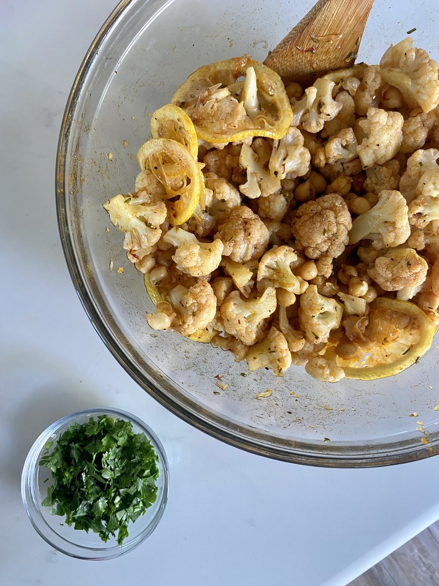 Closer view of the glass bowl of roasted cauliflower and chickpeas with lemon and a wooden spoon, chopped parsley in a small bowl