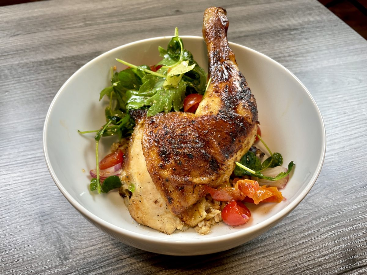 A crisp-skinned roasted chicken leg quarter standing in a wide white bowl over mixed greens, cherry tomatoes and sliced cucumber,