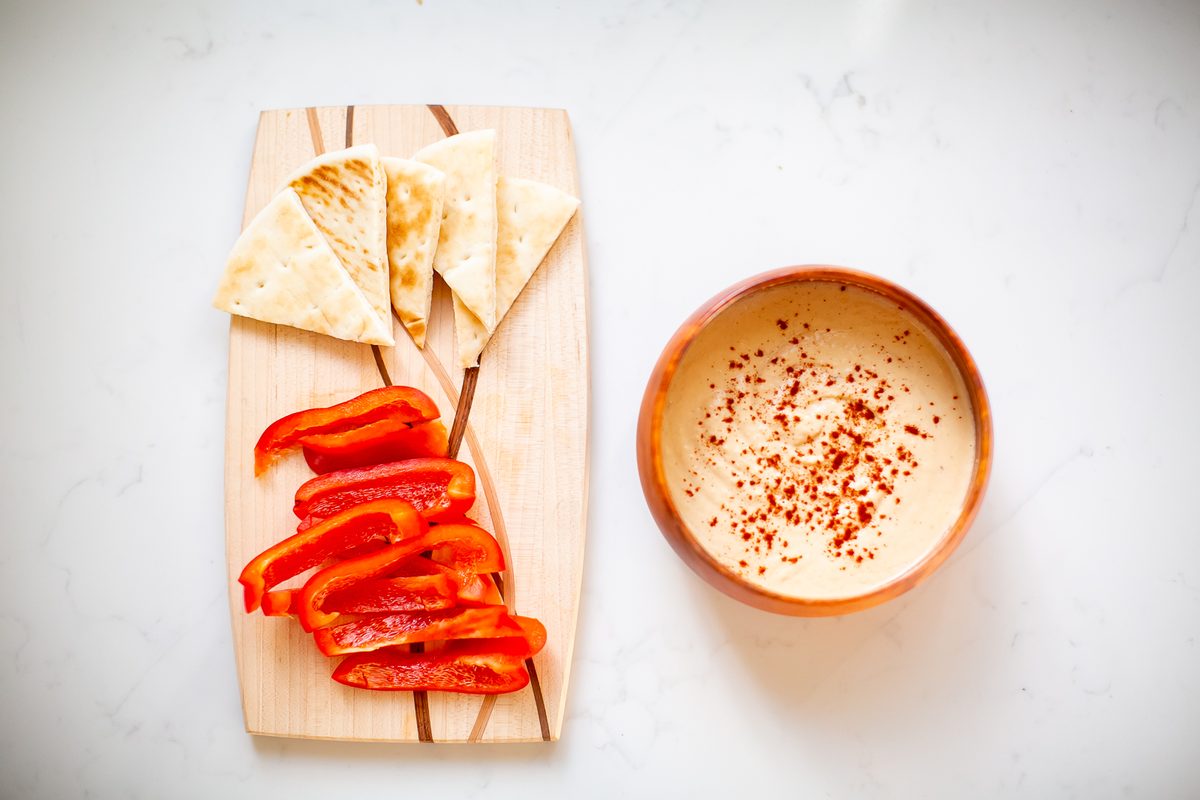 Overhead shot of a terracotta bowl of smooth hummus dusted with paprika beside a wooden board of toasted pita triangles and sliced