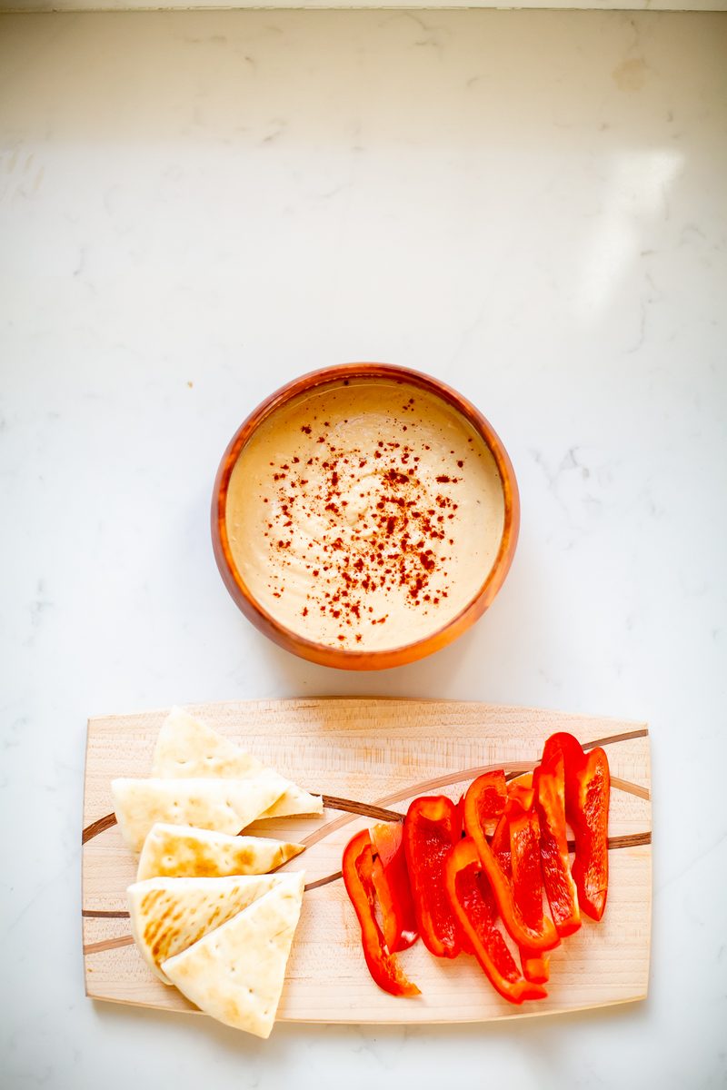Vertical overhead of a terracotta bowl of paprika-dusted hummus above a wooden board with pita wedges and red pepper strips on a w