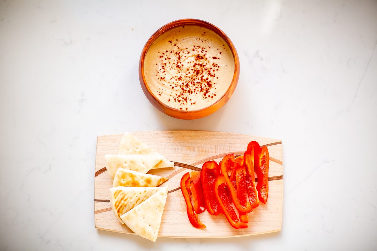 Overhead of a bowl of hummus with paprika next to a wooden serving board holding pita triangles and sliced red bell pepper, marble