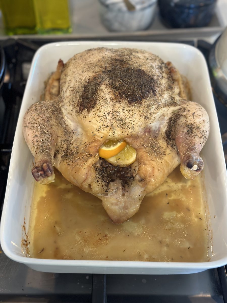A raw herb-rubbed whole chicken with a lemon slice tucked at its cavity, in a white square baking dish with pale liquid, on a stov