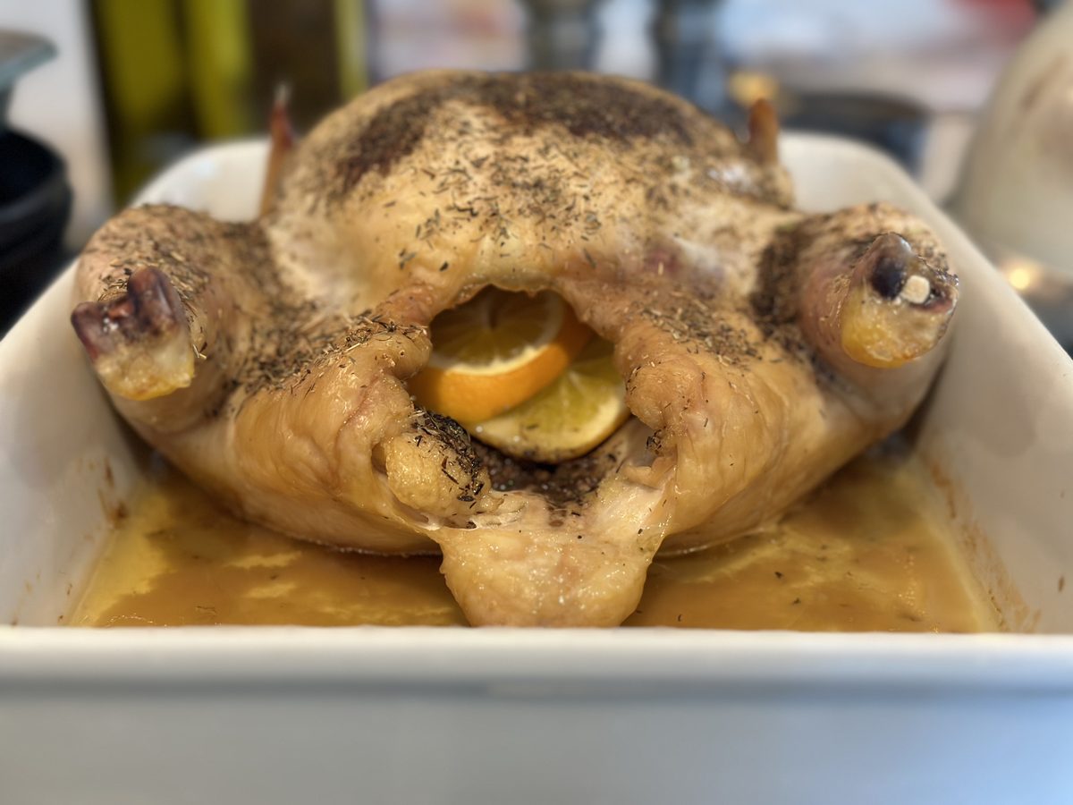A whole roasted chicken with browned herb-speckled skin and a lemon in the cavity, sitting in a white baking dish with pan juices.