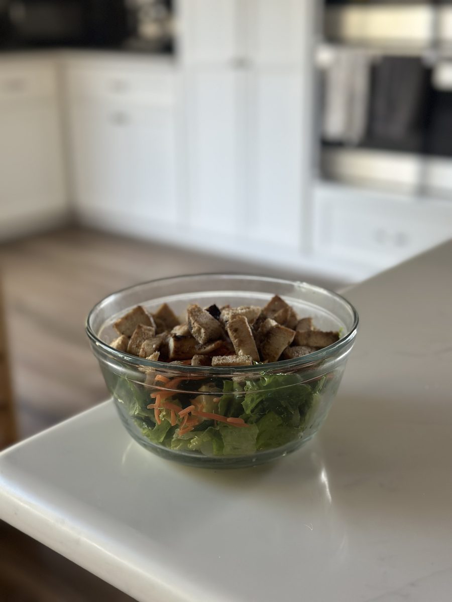A clear glass bowl of chopped green salad with shredded carrot, topped with a thick layer of cubed croutons, sitting alone on a wh