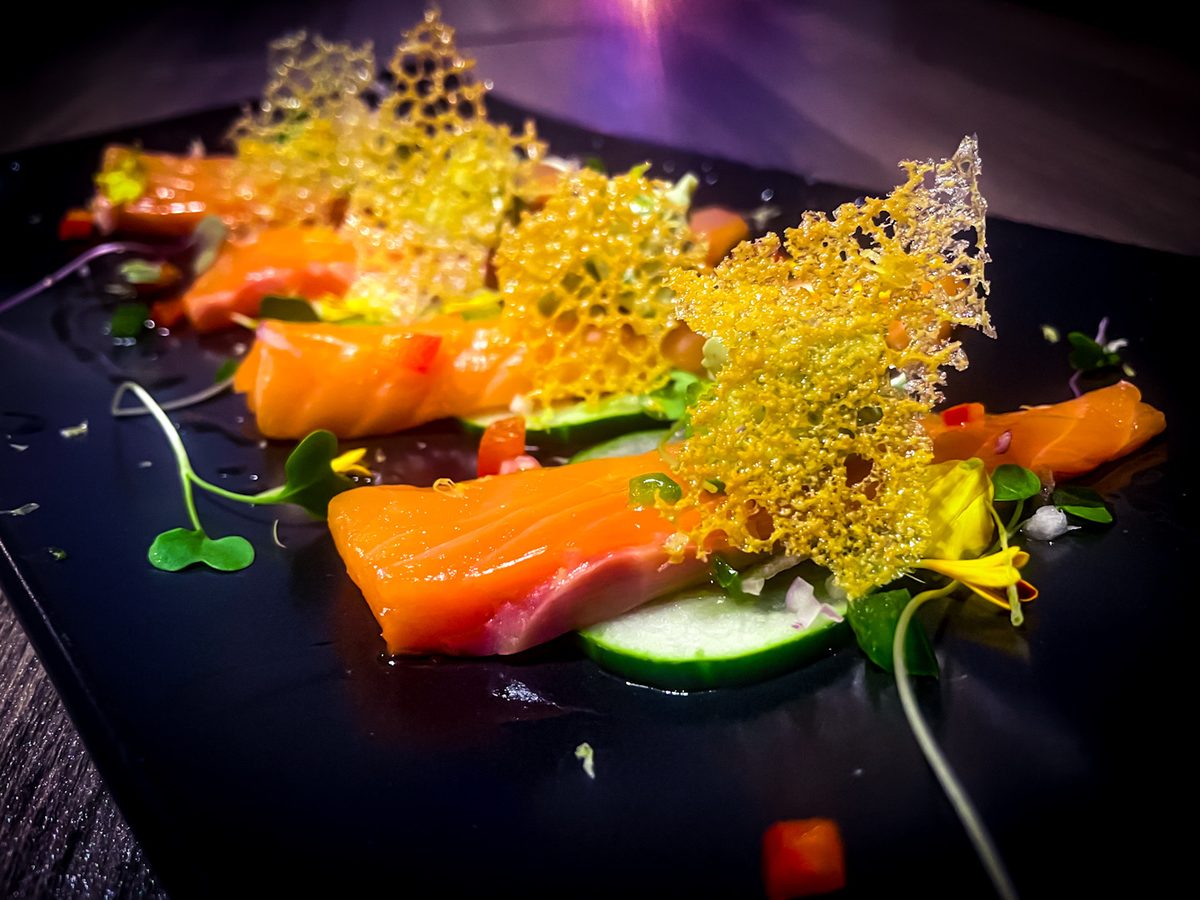 A dark slate plate with fine-dining plating of orange salmon slices, cucumber rounds, microgreens, dots of sauce and a golden spun